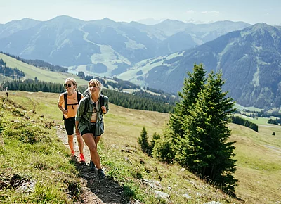 Zwei Frauen wandern bei strahlendem Sonnenschein auf einem alpinen Wanderweg in den Bergen rund um Hinterglemm, mit weitem Blick ins grüne Tal und auf umliegende Gipfel.
