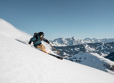 in Skifahrer in dynamischer Bewegung fährt durch unberührten Pulverschnee an einem sonnigen Wintertag in Saalbach-Hinterglemm. Im Hintergrund erstreckt sich eine beeindruckende Bergkulisse mit schneebedeckten Gipfeln unter klarem, blauem Himmel.